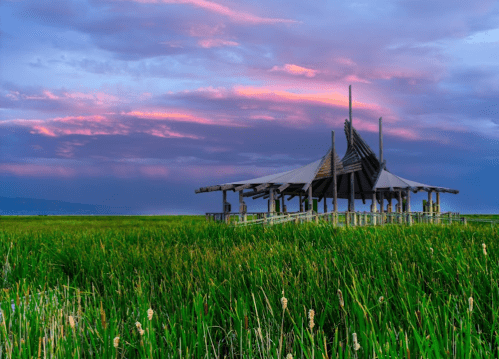 A wooden structure on stilts surrounded by tall grass, set against a colorful sunset sky.